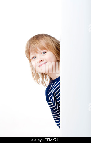 Curioso ragazzo con lunghi capelli biondi che spuntavano da dietro un pannello bianco sorridente Foto Stock