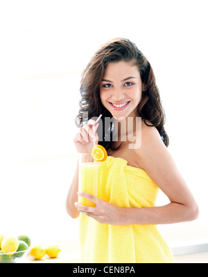 Femmina con ondulata lungo brunette capelli, avvolto in giallo asciugamano, tenendo in mano un bicchiere di succo di arancia, sorridente, guardando la fotocamera Foto Stock