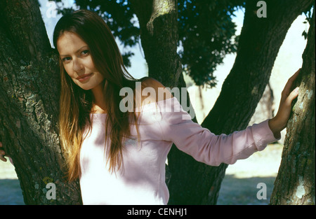 Bruna femmina diritta centro capelli di troncaggio indossando rosa off la spalla camicia a maniche lunghe in piedi un albero cercando Foto Stock