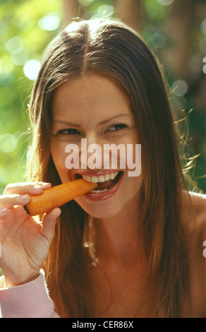 Bruna femmina diritta centro capelli di troncaggio e lucida labbra, mangiando una carota in un giardino, la bocca aperta Lei è sorridente e Foto Stock