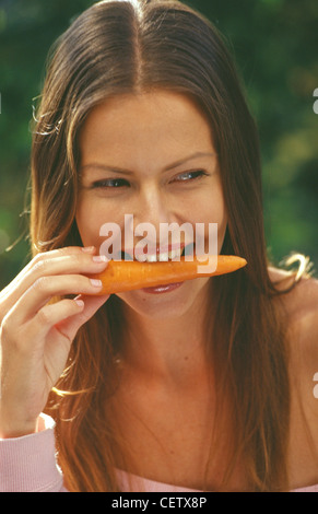 Bruna femmina diritta centro capelli di troncaggio e lucida labbra, mangiando una carota in un giardino lei è sempre sorridente e guardando lateralmente Foto Stock