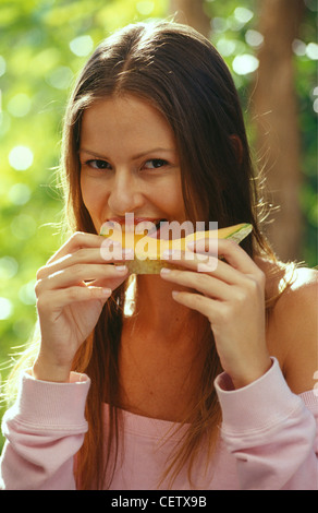 Bruna femmina diritta centro capelli di troncaggio, mangiare arancione di melone Honeydew entrambe le mani in un giardino indossa rosa off il Foto Stock