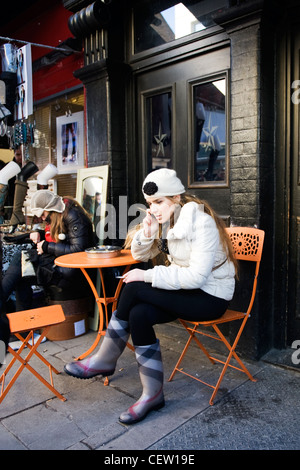 Portobello Road Street Market London elegante piuttosto giovane ragazza bionda abbigliamento invernale seduto telefono cellulare il fumo arancione sedia tavolo Foto Stock