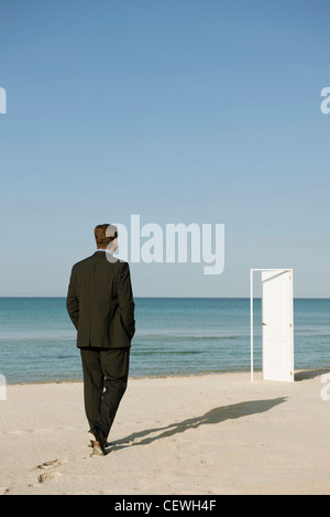 Imprenditore camminando sulla spiaggia verso la semi-porta aperta, vista posteriore Foto Stock