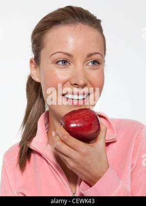 Femmina con capelli biondi legati a coda di cavallo, indossa una camicia rosa, mangiare una mela rossa, sorridente, guardando al lato Foto Stock