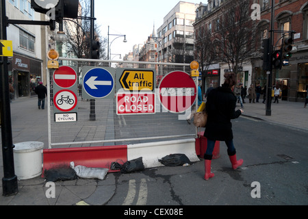 Una femmina di pedone indossare stivali rossi dodging barriera con segni il reindirizzamento del traffico su sezione chiusa di Oxford Street Londra Foto Stock