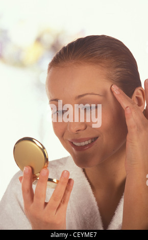Donna capelli castani off faccia bianca da indossare accappatoio in spugna guardando a se stessa in un compatto mirrtouching tempio dita sorridente Foto Stock