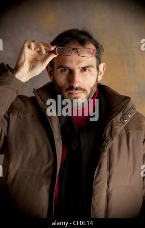 Giovane uomo che indossa cappotto marrone sollevamento suoi occhiali con mano destra studio shot illuminazione stroboscopica guardando la telecamera 20-30 30-40 caldo Foto Stock