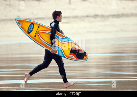 Un surfista in tutta Fistral Beach portando la sua tavola da surf. Foto Stock