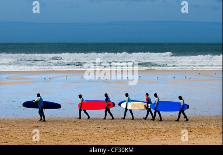 Surfers camminando in una linea che trasporta le loro tavole da surf attraverso Fistral Beach. Foto Stock