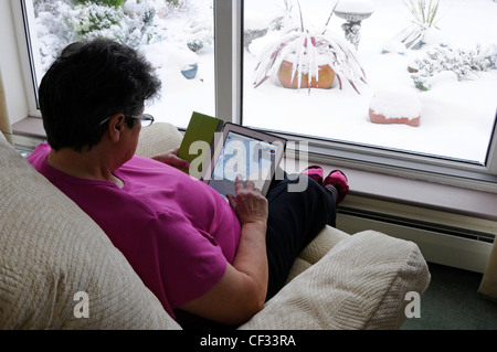 Donna a casa da sola che utilizza un tablet Apple iPad seduto poltrona calda al coperto da finestra scena invernale e neve pesante Indietro giardino Essex Inghilterra UK Foto Stock
