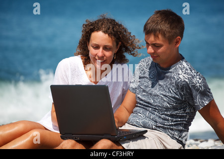 L uomo e la ragazza seduta sulla riva del mare. Con interesse, guardando il laptop in piedi sulle ginocchia. Close-up Foto Stock