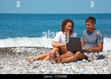 L uomo e la ragazza seduta sulla riva del mare. Con interesse, guardando il laptop in piedi sulle ginocchia. Foto Stock