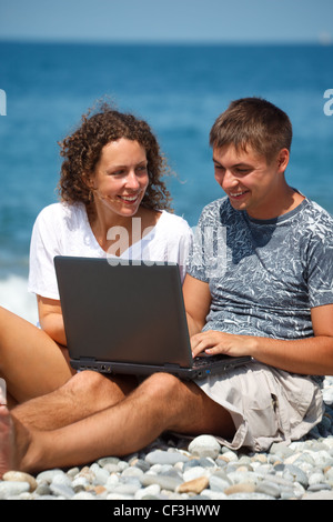 L uomo e la ragazza seduta sulla riva del mare. Con smile, guardando il laptop in piedi sulle ginocchia. Formato verticale Foto Stock