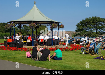Persone rilassante sotto il sole godendo di una banda di ottoni giocando nel palco per spettacoli in Crescent Gardens. Foto Stock