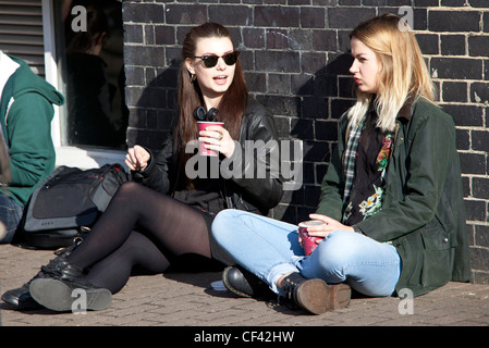 Ritratto di due giovani donne che parlano, bevono e siedono sul marciapiede, Londra, Inghilterra, Regno Unito. Foto Stock