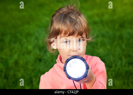 Ragazza che gioca con la lanterna. Ritratto di una ragazza contro un'erba. Sfondo dell'erba. bambina guarda nella lente. ragazza brilla una lanterna nella lente Foto Stock