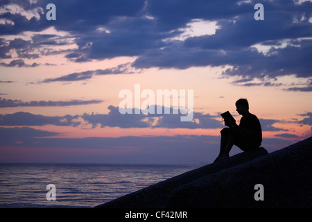 Silhouette Ragazzo seduto sulla scogliera in sera vicino al mare, legge prenota Foto Stock