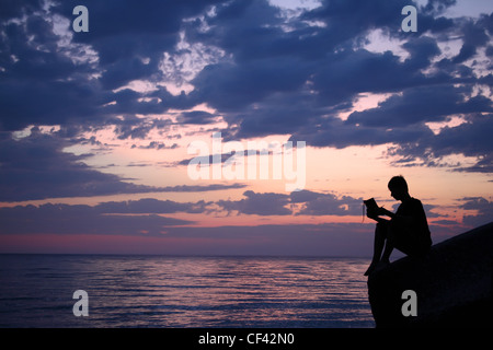 Silhouette Ragazzo seduto sulla scogliera in sera vicino al mare, legge prenota Foto Stock