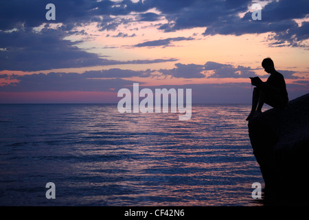 Silhouette Ragazzo seduto sulla scogliera in sera vicino al mare, legge prenota, ampio angolo Foto Stock