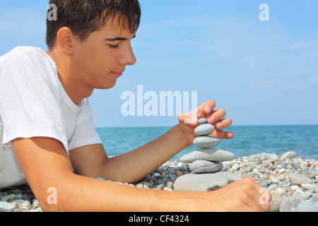 Ragazzo adolescente giacente su terreni sassosi seacoast, crea la piramide da ciottoli, guardando a piramide Foto Stock