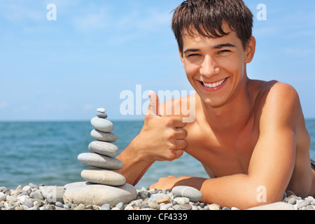 Ragazzo adolescente giacente su terreni sassosi seacoast, crea la piramide da ciottoli, mostra ок gesto Foto Stock