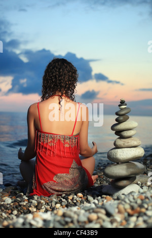 Bella donna in rosso sundress meditando vicino a piramide dal ciottolo sul litorale di pietra in serata, seduta dal retro Foto Stock