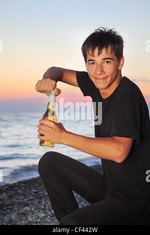 Ragazzo adolescente apre la birra imbottigliatore sul litorale di pietra in sera Foto Stock