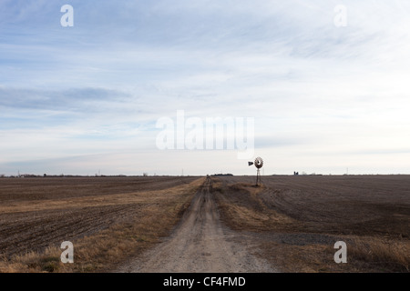 Un solitario strada sterrata in rural Illinois con un mulino a vento organizzandosi sopra i campi di colore marrone. Foto Stock