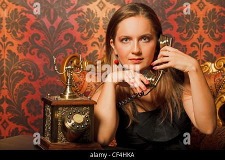 Donna in abito nero seduto su una sedia vintage e parlando al telefono vecchio. guarda nella telecamera Foto Stock