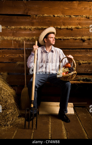 Giovane con forcone,con cesto di frutta e nel cappello di paglia seduta sul banco in legno capanna di registro Foto Stock