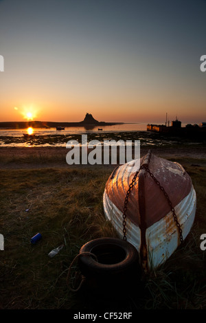 Una piccola barca capovolta sulla riva con Lindisfarne Castle profilarsi all alba la luce del sole in background. Foto Stock