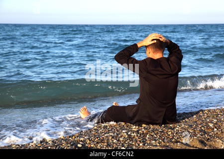 Intendo nudi uomo in tuta si siede indietro sulla costa di pietra e mette la mano sulla testa alla sera Foto Stock