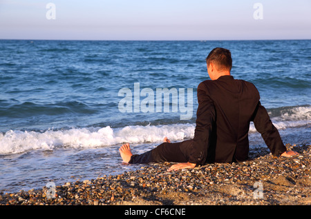 Nudi uomo nel business suit si siede indietro su pietra costa alla sera Foto Stock