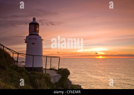 Polperro faro in primo piano come il sole sorge sopra l'orizzonte la creazione di un cielo rosa. Foto Stock