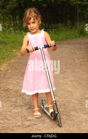 Bellissima bambina in abito rosa rides scooter sulla natura. foresta potrebbe essere visto in background Foto Stock