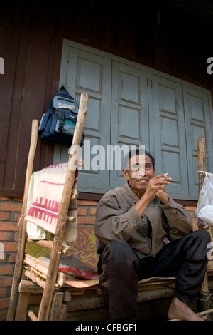 Un centro di età compresa tra uomo asiatico è seduta su una panchina di fumare una sigaretta in una casa a Luang Namtha, Laos. Foto Stock