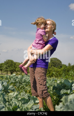 Multi-generazionale ritratto di donna anziana azienda piccola ragazza bionda con pig-tail in piena luce solare in un cavolo cappuccio patch in fattoria. Foto Stock