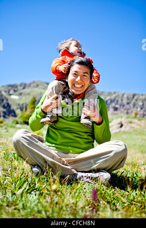 Una donna sorrisi come siede zampe trasversale in un prato con fiori selvaggi facendo un Buddha pongono. Suo figlio, un bambino di 2 anni, crawl Foto Stock