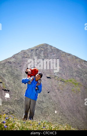 Una madre escursioni con il suo bambino di 2 anni attraverso un prato alpino che è disseminato di giallo fiori selvatici alpini (daisy simili) con tr Foto Stock