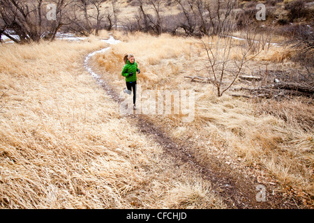 Una giovane donna corre un sentiero verso il basso attraverso l'erba Morta appena a nord di Dixon serbatoio, in un freddo giorno di un verde down jacket. Foto Stock