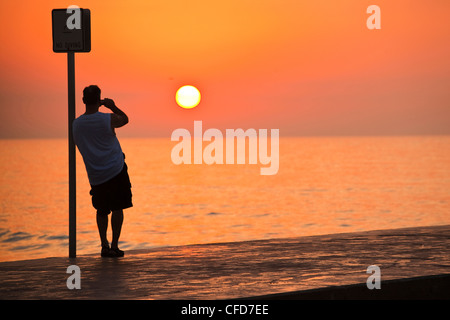 Uomo appoggiato a un segno posto e fotografare il sorgere del sole, Key West, Florida, Stati Uniti d'America. Foto Stock
