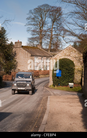 Vista frontale del Land Rover Defender in viaggio o durante la marcia su strada di campagna passato pittoreschi cottage - Bolton Abbey village, North Yorkshire, Inghilterra, Regno Unito. Foto Stock