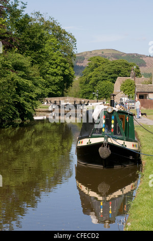 Barca stretta ormeggiata vicino a blocco a lato del panoramico canale rurale Leeds Liverpool e riflesso in acqua il giorno di sole - Gargrave, North Yorkshire, Inghilterra, Regno Unito. Foto Stock