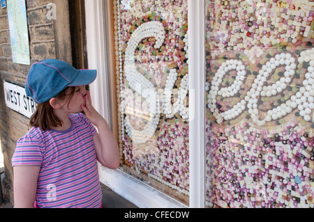 Un giovane bambino (in sun hat) guardando stupito (mano sulla bocca) a allettante display artistico di squisiti dolci di riempimento nella finestra shop - Gargrave, nello Yorkshire, Regno Unito. Foto Stock