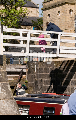 2 persone su passerella (donna e bambina) guardano in basso come lady su narrowboat passa al di sotto di esse - Aumento di cinque serrature, Bingley, Leeds Liverpool Canal, Inghilterra. Foto Stock