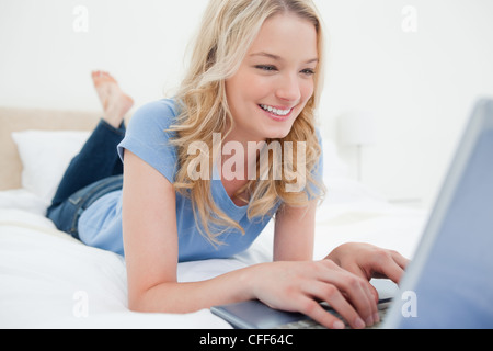 Donna sdraiata sul letto digitando nel suo computer portatile e sorridente Foto Stock