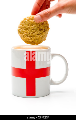 Una mano caucasica che tiene un biscotto pronto per essere infilato in una tazza bianca con una bandiera inglese sul suo lato, riempita di tè. Foto Stock