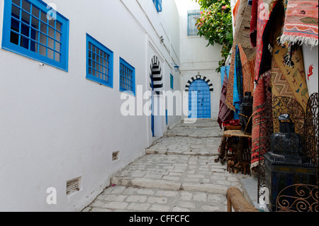 Sidi Bou Said. La Tunisia. Vista dei tappeti di souvenir in vendita lungo stretti vicoli in ciottoli di bianco e nero con blu arcuata in corrispondenza della porta Foto Stock