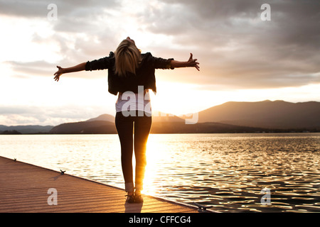Una giovane e bella donna che guarda nella distanza mantiene le sue braccia fuori abbracciando il tramonto su un lago in Idaho. Foto Stock